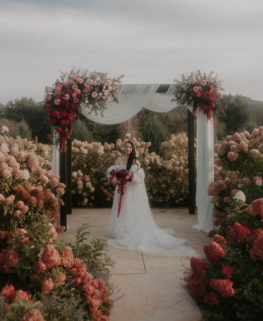 A bride standing beneath an arbor decorated with floral installations in pink and red.
