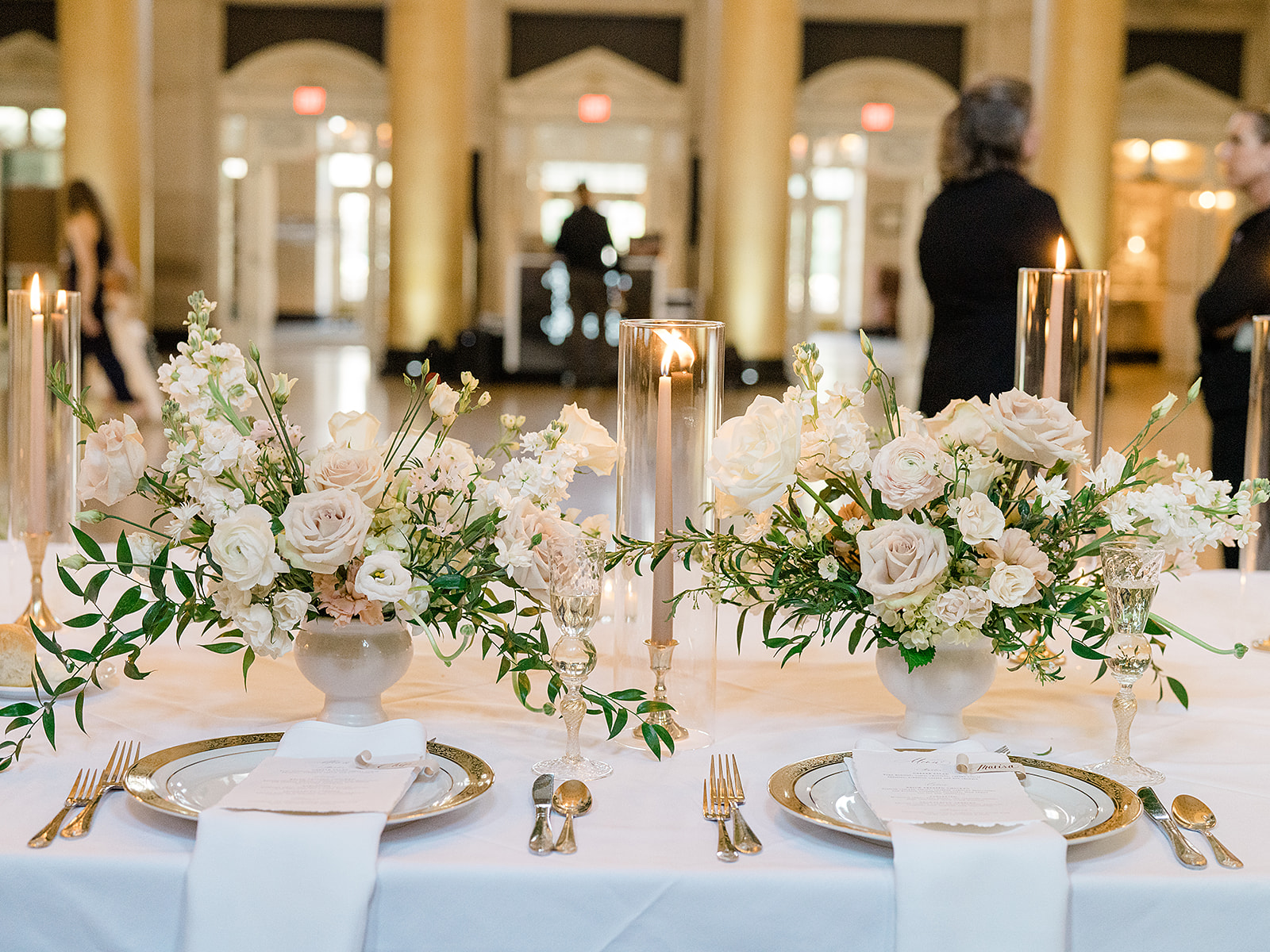 Wedding floral centerpieces in white pedestal vases on guest table with taper candles.