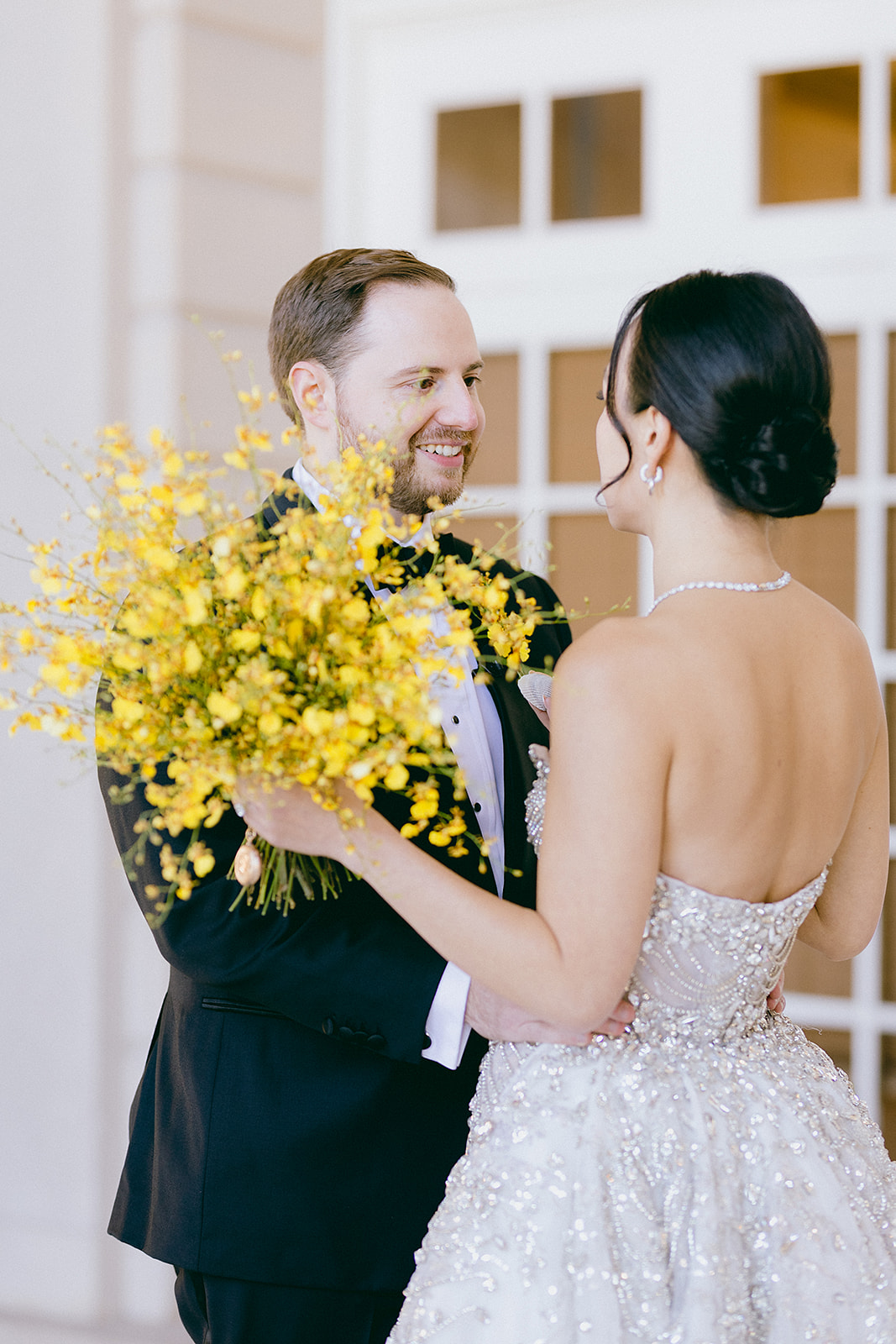 A smiling couple shares a romantic moment, with the bride holding a vibrant bouquet of acid yellow oncidium orchids. She wears a sparkling silver gown, and the groom is dressed in a classic tuxedo, surrounded by a soft indoor setting.
