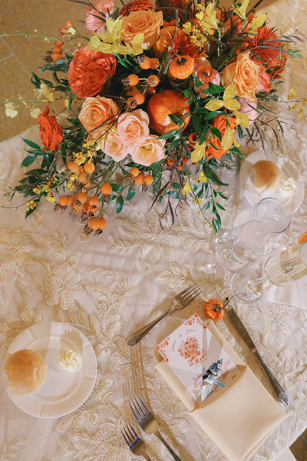 A beautifully arranged fall-themed centerpiece featuring orange and pink roses, acid yellow oncidium orchids, pumpkins, and mixed berries, set on an elegant lace tablecloth, complemented by plates, cutlery, and glasses.