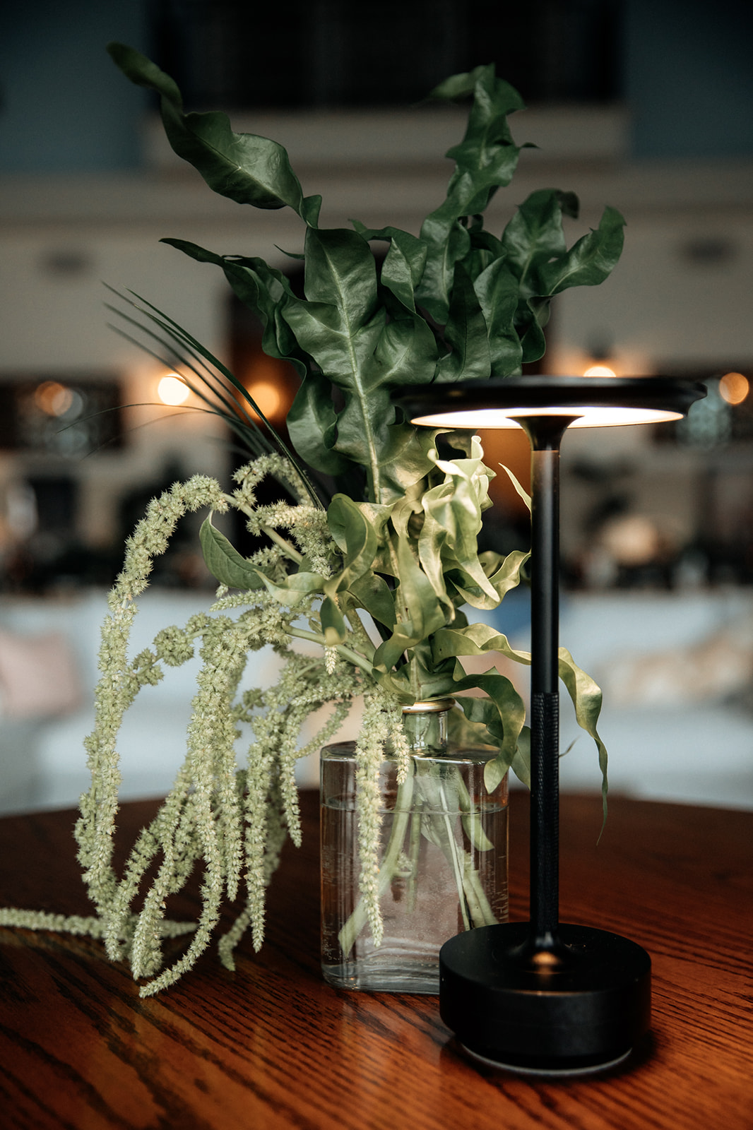 A close-up of a glass vase filled with tropical foliage, including large green leaves and delicate light green flowers, complemented by a modern black lamp on a wooden table.