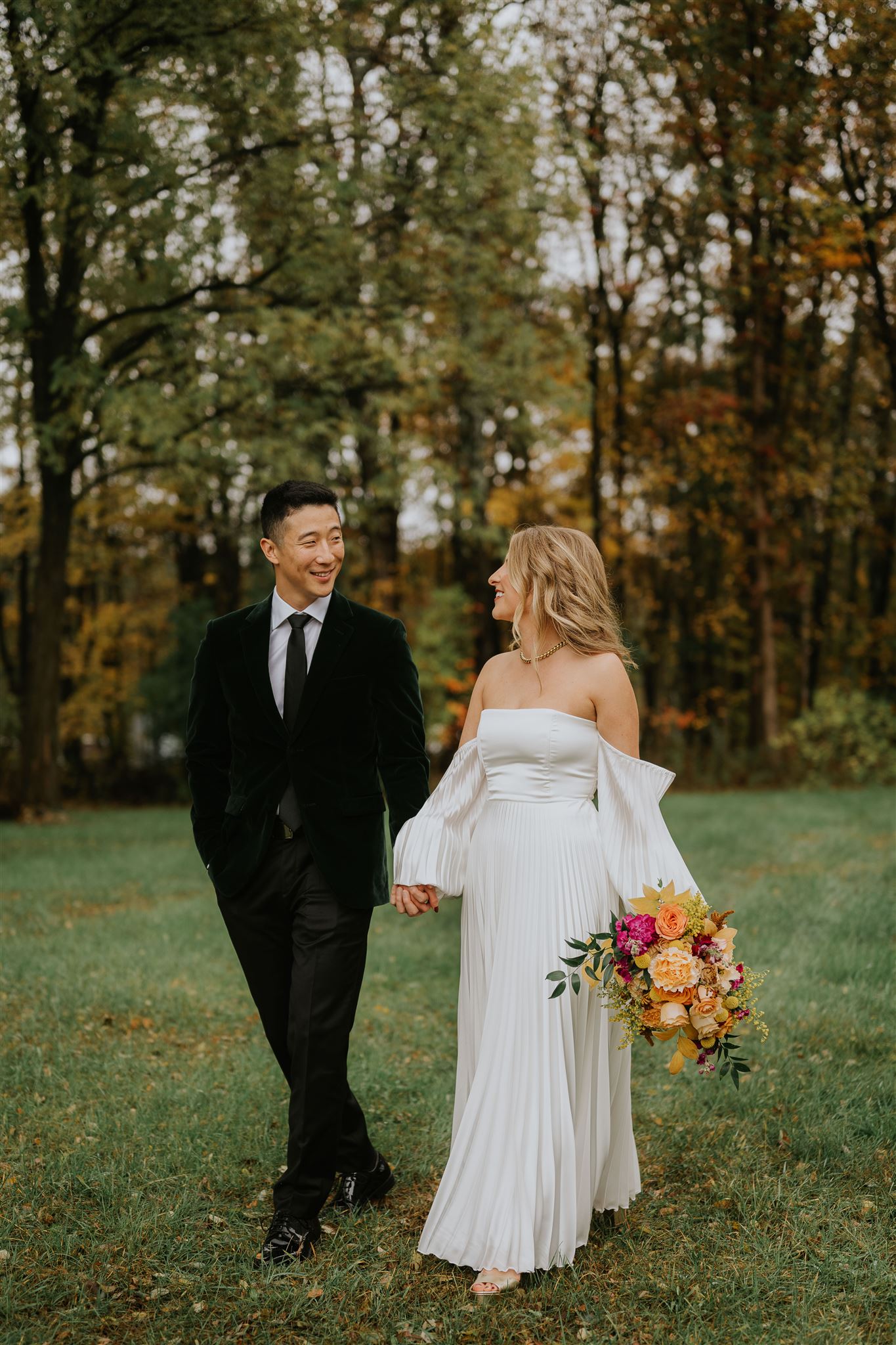 A bride and groom walk holding hands in front of a forest on an autumn day.
