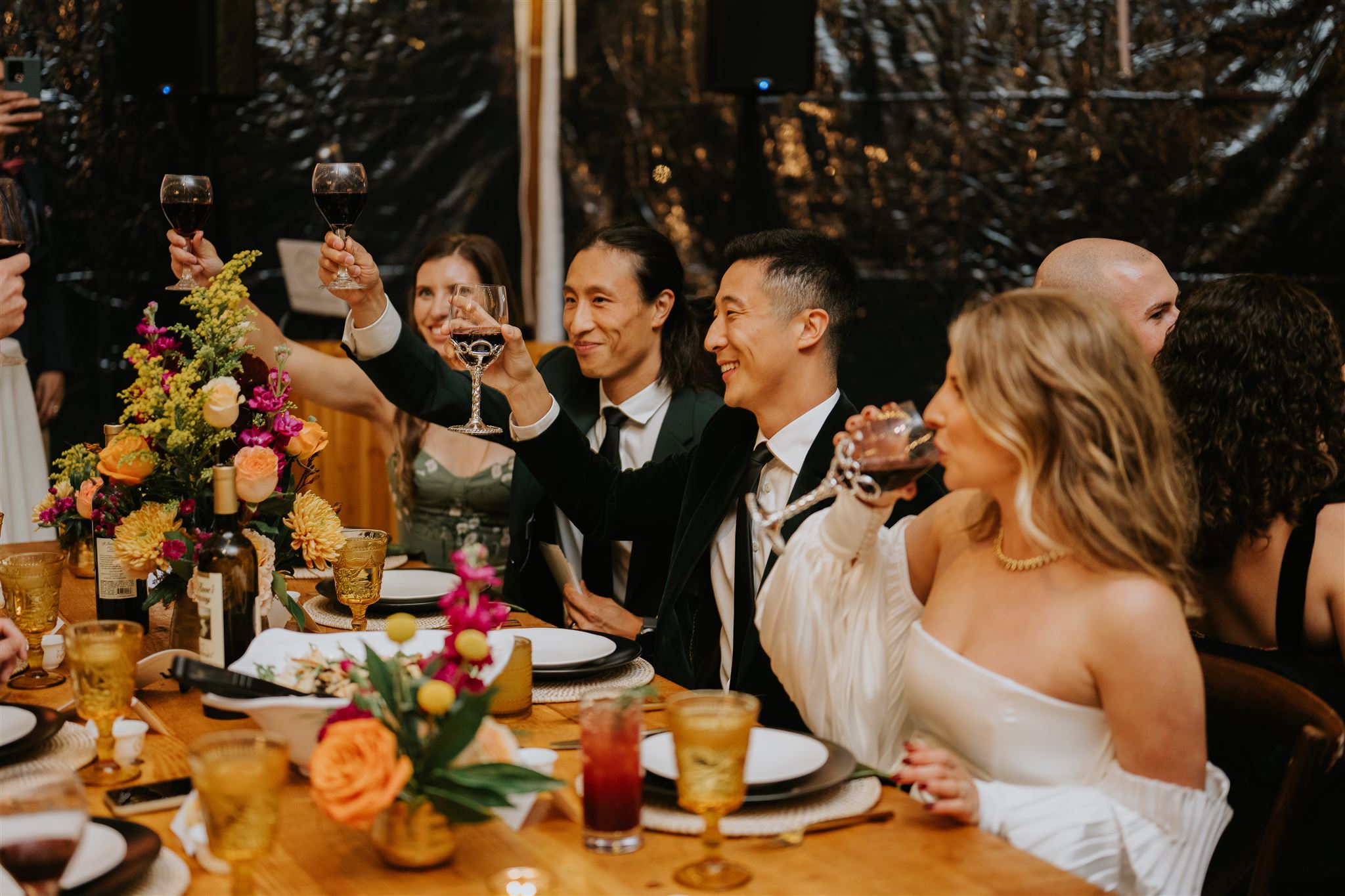 A bride and groom laughing with friends at a table with floral arrangements.