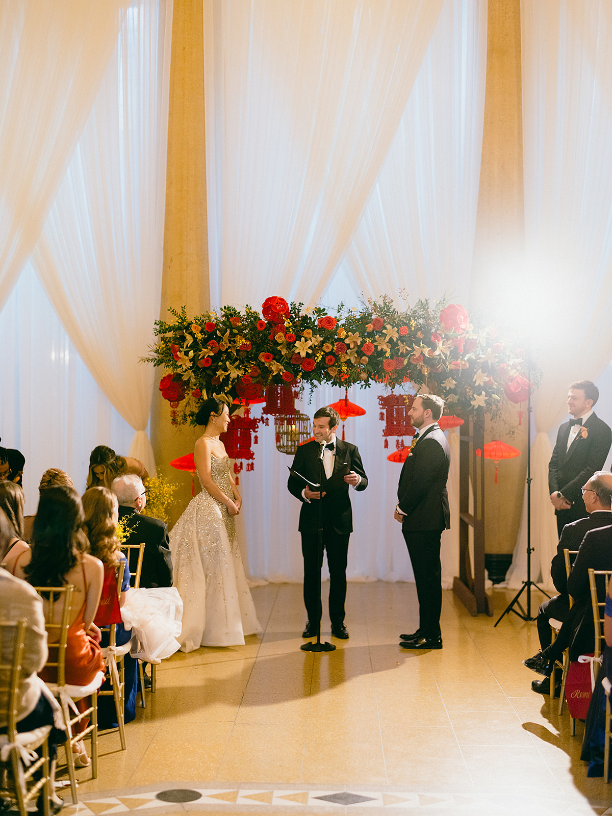 A bride and groom stand at the altar during their wedding ceremony, surrounded by guests. The altar features a vibrant floral arrangement and festive lanterns, creating a celebratory atmosphere.