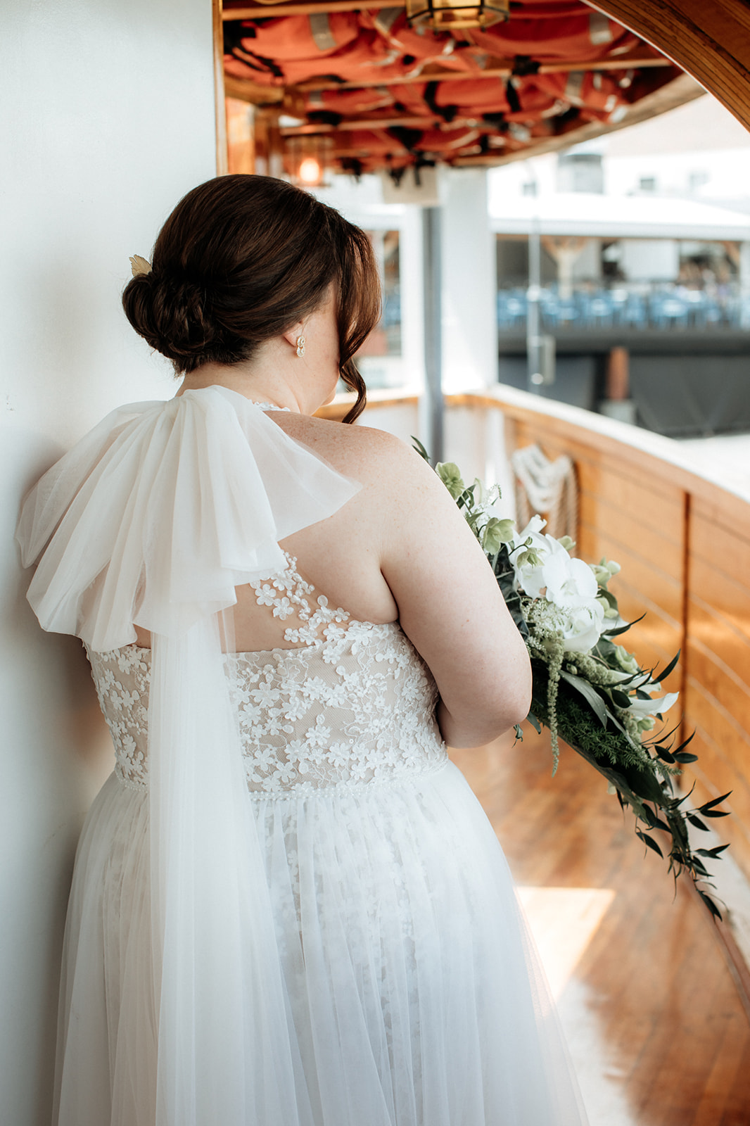 A bride in a lace-back wedding dress with a flowing veil stands on a wooden deck, holding a bouquet of white flowers and foliage, looking away from the camera.